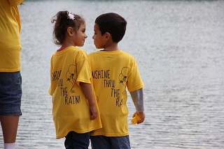 Two children wearing Sunshine Thru the Rain shirts stand together beside the water